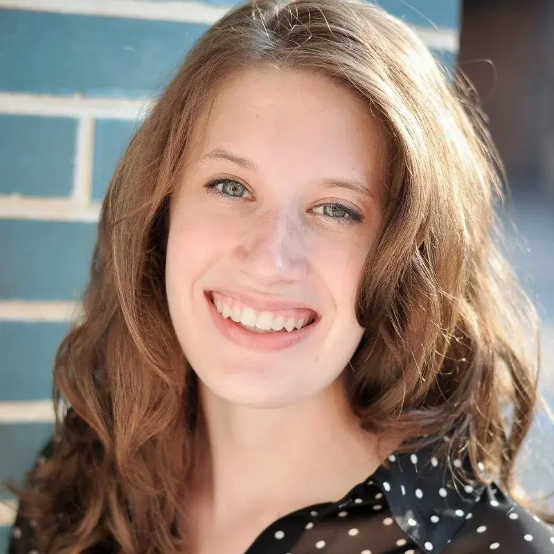 A young woman with long, wavy auburn hair, smiling, outdoors near a blue brick wall.